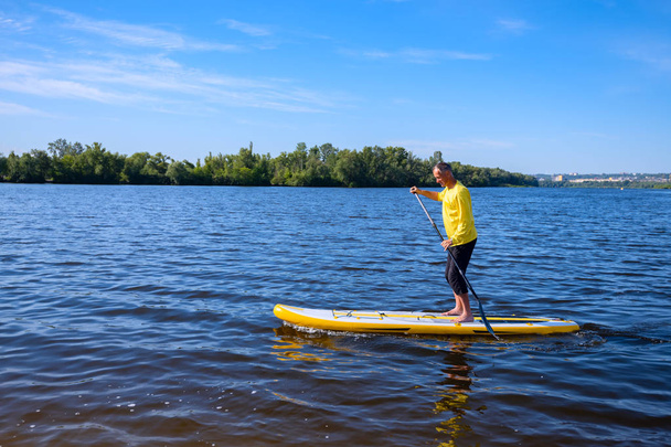 Happy adult man is training to sail on a SUP board on sunny morning on cityscape background. Stand up paddle boarding - awesome active recreation during vacation. - Fotó, kép