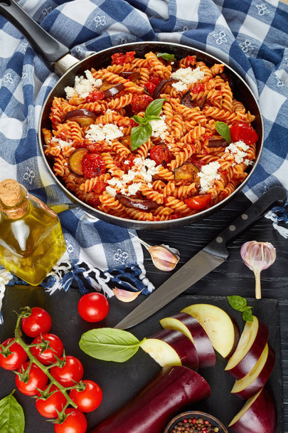 Vegetarian Italian Pasta fusilli alla Norma with eggplant, tomatoes, basil, ricotta cheese in a skillet on a wooden table with ingredients at the background, vertical view from above, close-up - Photo, image