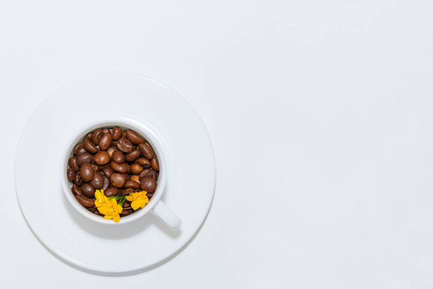 white cup and saucer on a white background top view. in a cup are poured coffee beans and yellow flowers - Photo, Image