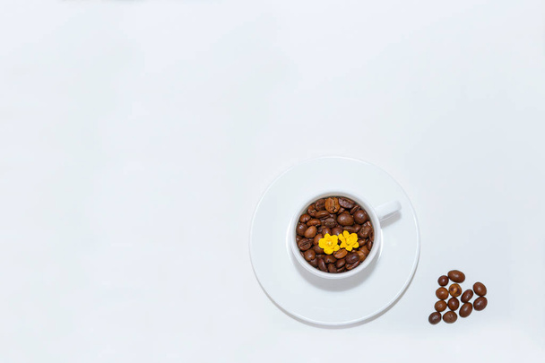 白いカップとソーサー白い背景の上に表示します。カップにコーヒー豆と黄色の花を注いだが。コーヒー豆の白い背景の心に - 写真・画像