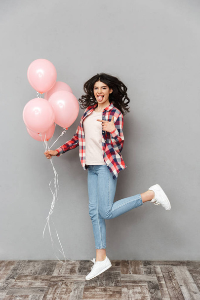 Image of cute beautiful young lady jumping isolated over grey background wall holding balloons pointing. - 写真・画像