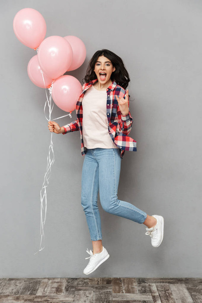 Image of cute beautiful young lady jumping isolated over grey background wall holding balloons make rock gesture. - Foto, immagini
