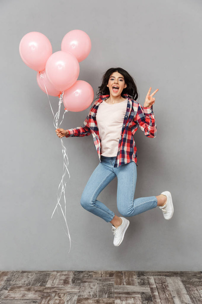 Image of cute beautiful young lady jumping isolated over grey background wall holding balloons make peace gesture. - Foto, afbeelding
