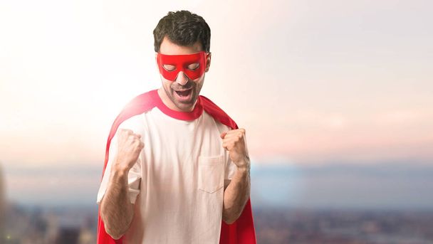 Супергерой в маске и красном плаще празднует победу и радуется тому, что выиграл приз на фоне заката
 - Фото, изображение