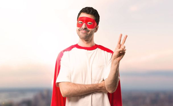 Homem super-herói com máscara e capa vermelha sorrindo e mostrando sinal de vitória com um rosto alegre em um fundo de pôr do sol
 - Foto, Imagem