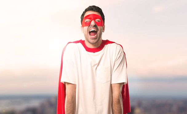 Superheld man met masker en rode cape schreeuwen aan de voorkant met de mond wijd open op de achtergrond van een zonsondergang - Foto, afbeelding