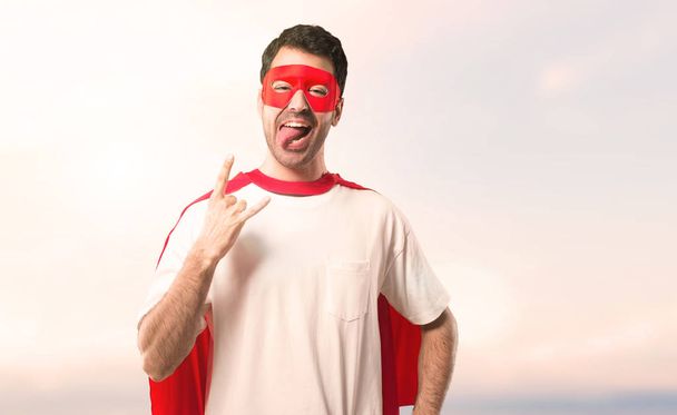 Hombre superhéroe con máscara y capa roja mostrando la lengua en la cámara teniendo mirada divertida y sacando los cuernos sobre un fondo de puesta de sol
 - Foto, imagen