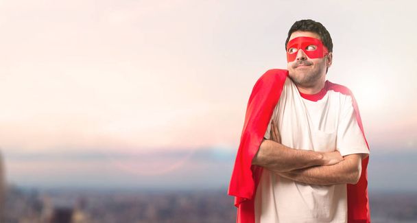 Superhero man with mask and red cape making unimportant and doubts gesture while lifting the shoulders on a sunset background - Photo, Image