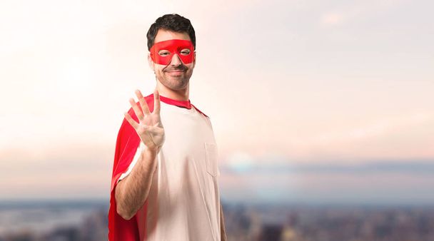 Superhelden-Mann mit Maske und rotem Umhang glücklich und zählt mit den Fingern vier vor Sonnenuntergang - Foto, Bild