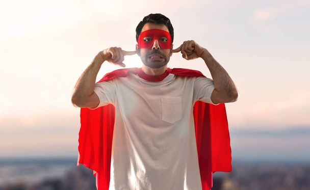 Homem super-herói com máscara e capa vermelha cobrindo seus ouvidos em um fundo pôr do sol
 - Foto, Imagem