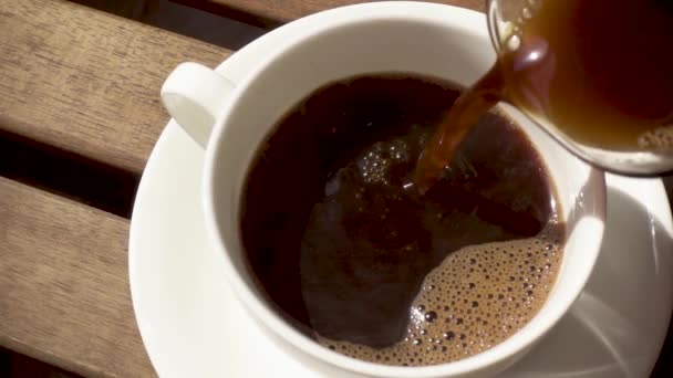 heißer schwarzer Kaffee aus der Kaffeekanne in eine weiße Porzellantasse auf einer Untertasse auf einem Holztisch in Zeitlupe gießen - Filmmaterial, Video