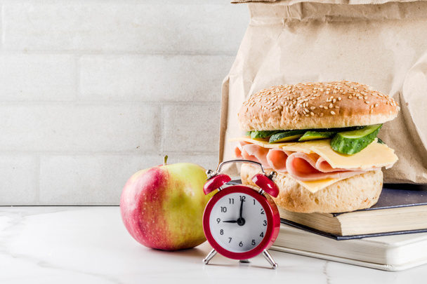 Концепція здорової шкільної їжі, паперовий мішок з обідом, яблуком, сендвічем, книгами та будильником на білому кухонному столі копіювання простору
 - Фото, зображення