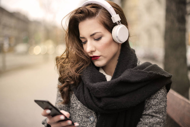 Νεαρή όμορφη γυναίκα με ακουστικά τον έλεγχο smartphone της στην πόλη. - Φωτογραφία, εικόνα