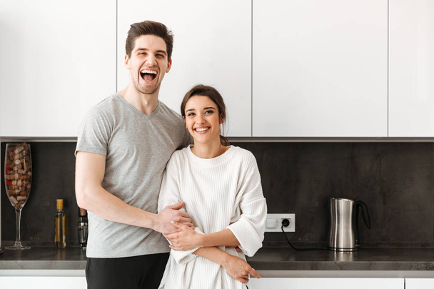 Portrait of a cheerful young couple hugging while standing at the kitchen at home - Foto, afbeelding