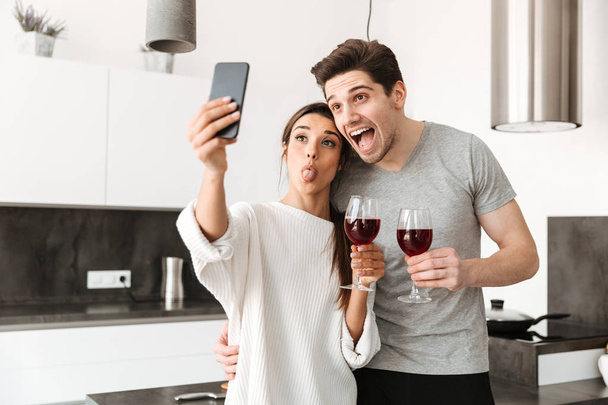 Porträt eines glücklichen jungen Paares, das ein Selfie macht, während es in der Küche mit Weingläsern steht - Foto, Bild