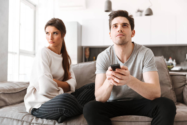 Portrait of an angry young woman sitting with her boyfriend on a couch while he is watching TV - Photo, image