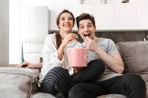 Portrait of a smiling young couple relaxing on a couch at home while watching TV and eating popcorn - Photo, image