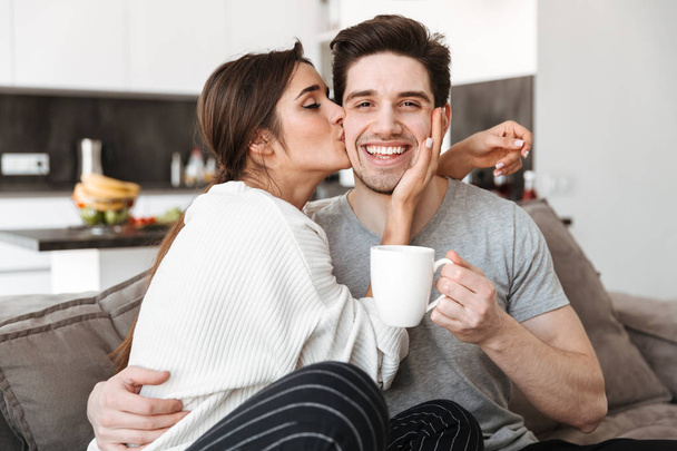 Portrait of a happy young couple drinking coffee while sitting on a couch and kissing - Foto, afbeelding