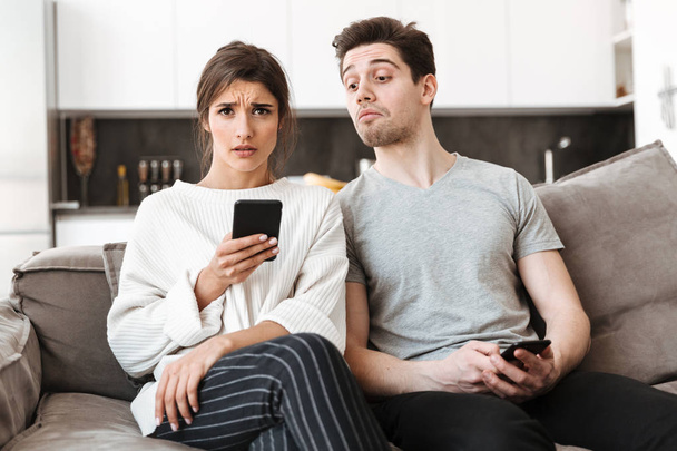 Portrait of a n upset young woman holding mobile phone while sitting on a couch with her boyfriend at home - Photo, Image