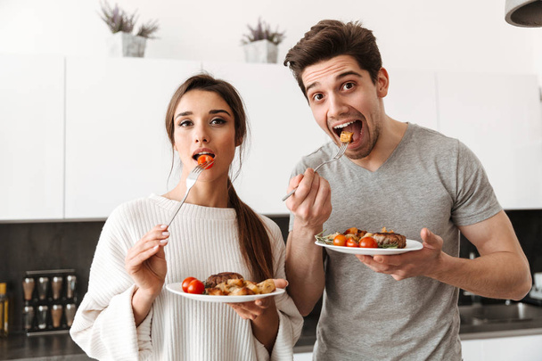 Portrait of a delighted young couple holding dinner plates with food while standing at the kitchen - Foto, imagen