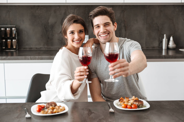 Portrait of a happy young couple holding glasses of wine while sitting at the kitchen table and having dinner - Фото, зображення