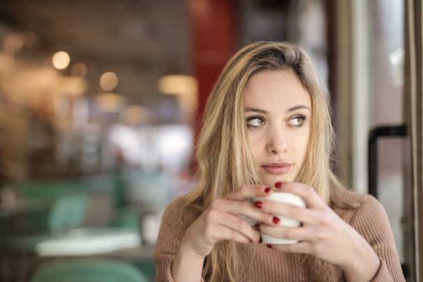 Ξανθιά νεαρή γυναίκα σε ένα bistro κοιτάζει έξω από το παράθυρο πίνοντας καφέ. - Φωτογραφία, εικόνα