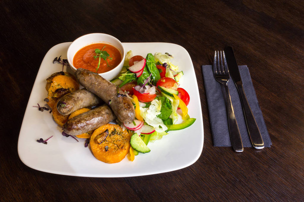 Смачні смажені ковбаски та овочевий салат у білій страві з червоним соусом на коричневому столі з виделкою та ножем
 - Фото, зображення