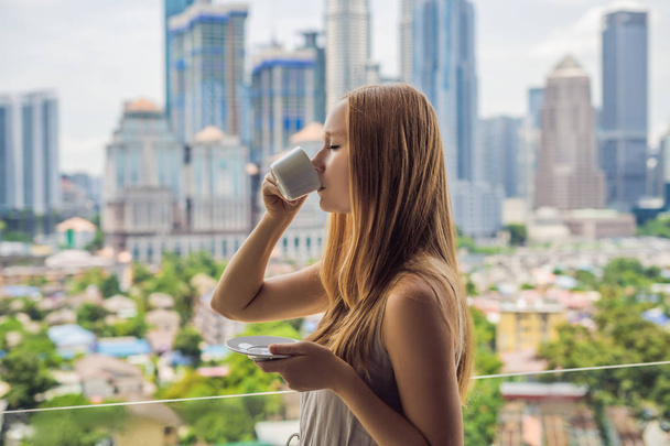Young woman drinks coffee in the morning on the balcony overlooking the big city and skyscrapers. - Foto, afbeelding