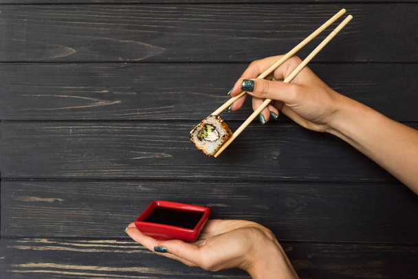 hands holding chopsticks with sushi and soy sauce in a red bowl on a black wooden background - Foto, Imagem