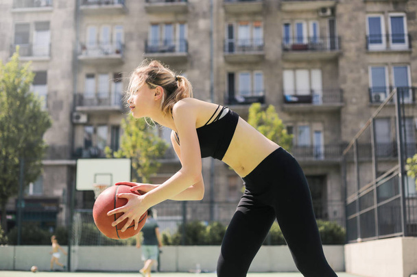 Young blonde teenager in fitness outfit playing basketball in the city. - Фото, зображення
