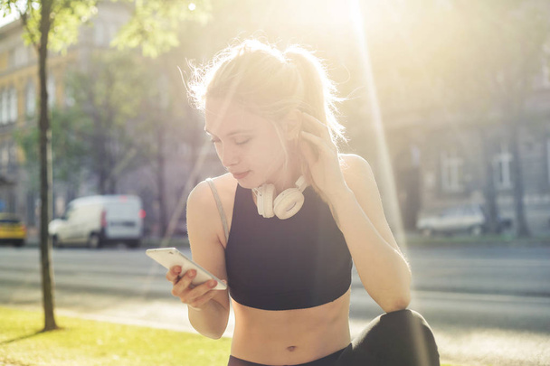 Young blonde woman in fitness outfit with headphones checking her smartphone in the city. - Fotó, kép