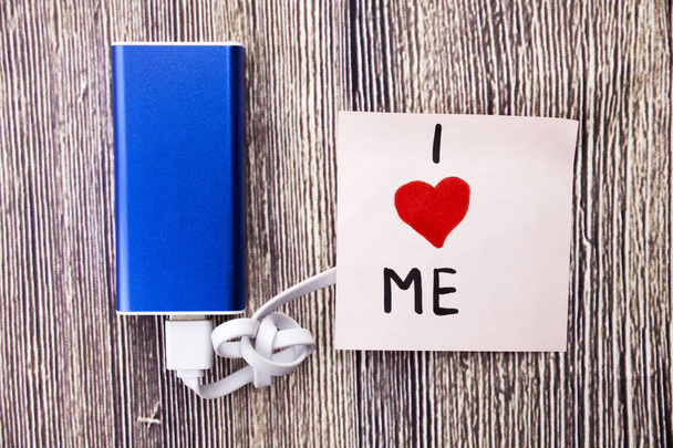 I love me heart handwritten on note paper in different colors and heart shape in red color. Energy storage device of blue color with cable on wooden background. selfish or self love Concept. - Photo, Image