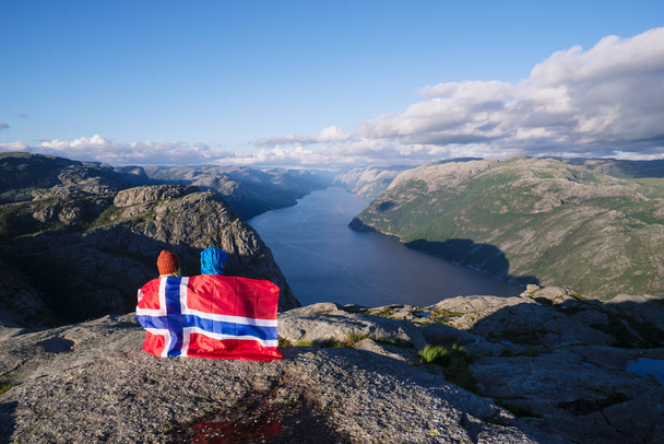 Preikestolen pathway. Couple with the flag of Norway looks at the panorama of the Lysefjord. Tourist attraction. Sunny weather in the mountains - Photo, Image