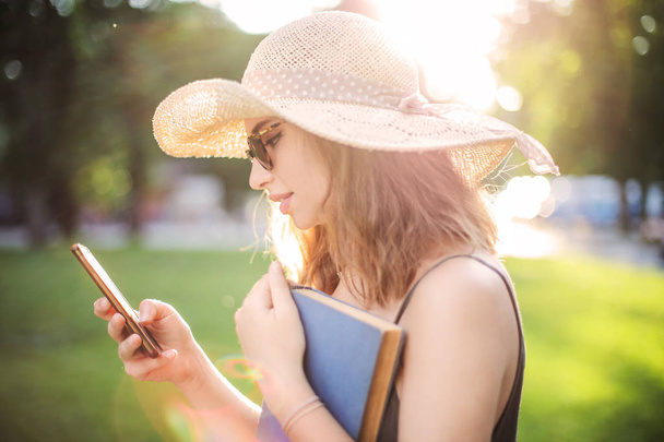 Young teenager wearing a straw hat and sunglasses checking her smartphone in a park outdoor on a summer day. - Foto, Bild
