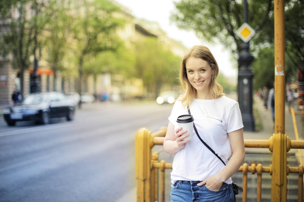 Νεαρή ξανθιά γυναίκα στέκεται στο δρόμο με ένα καφέ-to-go και χαμογελαστός θετικά. - Φωτογραφία, εικόνα