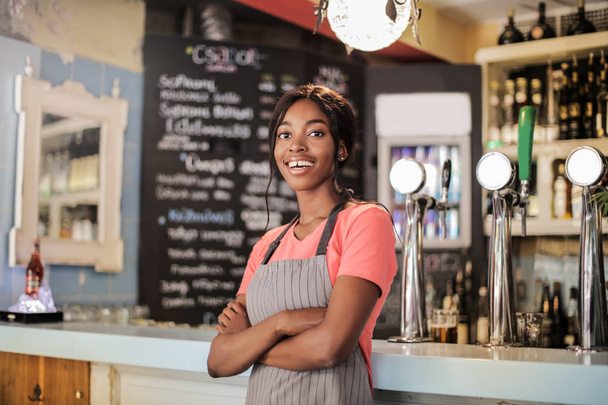 Young beautiful Afro woman as waitress standing in front of the bar and smiling. - Fotó, kép