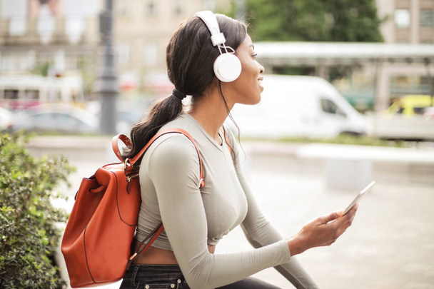 Young beautiful Afro woman with headphone and backpack sitting and waiting on the street with her smartphone in her hand. - Foto, afbeelding