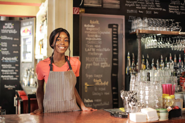 Young beautiful Afro waitress standing and smiling behind the countertop in a bistro. - Fotó, kép