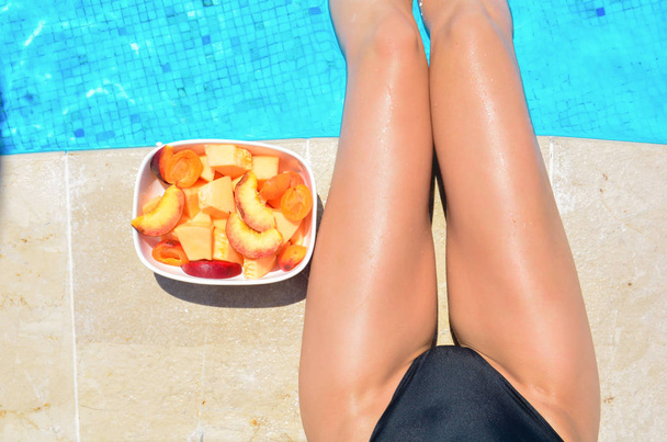 日焼けした美脚。リラックスして果物を食べる若い女性。薄い細身のフィットの女の子の足の近くのプールでボウルにフルーツ。フルーツ サラダ、健康食品。夏のコンセプト - 写真・画像