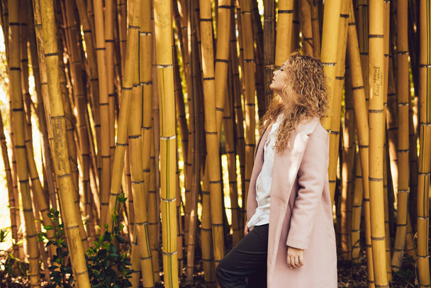 stylish young woman in trendy pink coat posing in park next to bamboo trees - Fotó, kép