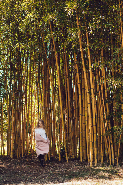 fashionable stylish woman in a pink coat walking in the park, posing next to tall bamboo trees - Foto, afbeelding