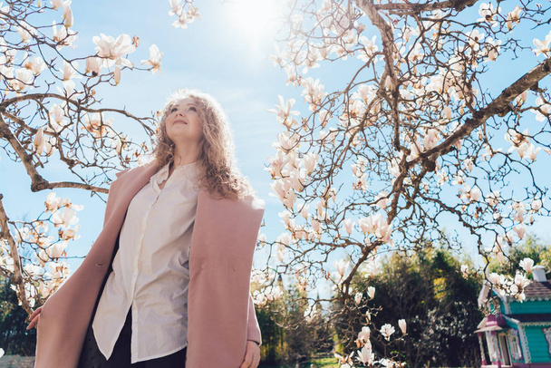 stylish elegant woman in a pink coat posing under a blue sky next to a blooming magnolia - Foto, afbeelding