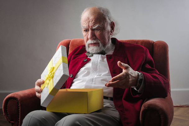Old man with white beard and grey hair looking surprised while opening a gift. - Valokuva, kuva