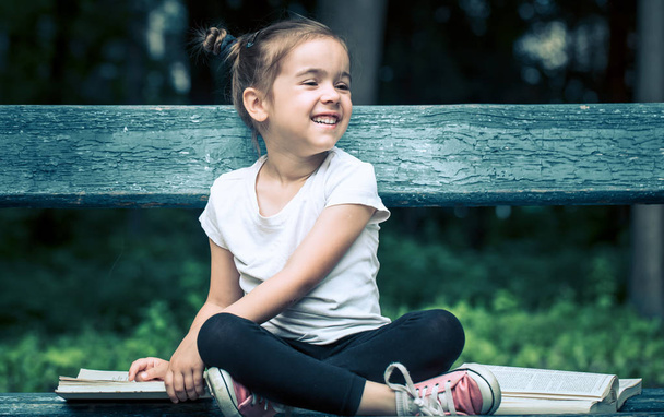 το μικρό χαριτωμένο κορίτσι που κάθεται σε ένα παγκάκι και διαβάζοντας ένα βιβλίο στο πάρκο σε φόντο πράσινο της φύσης - Φωτογραφία, εικόνα