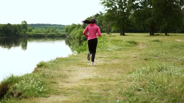 A lány rózsaszín kabátot és fekete nadrágot fut a maratoni felkészülés fejhallgató-folyó közelében - Felvétel, videó