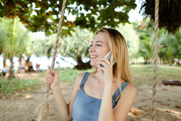 Joven mujer agradable montar swing y hablar por smatrphone, arena y árbol en el fondo
. - Foto, Imagen