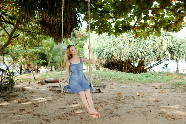 Young cute female person wearing jeans dress and riding on swing, sand in background. - Foto, imagen