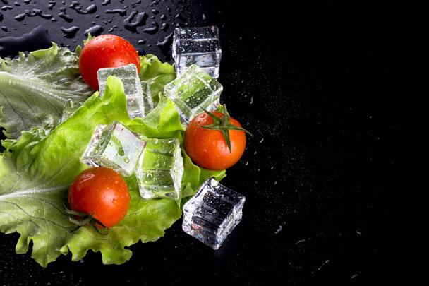 Banch of red cherry tomatos, green salad and ice cubes on black wet table. Selective focus. - 写真・画像