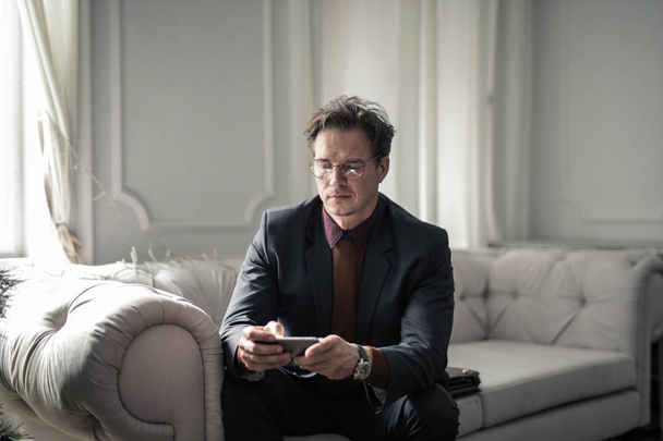 Businessman with glasses sitting on a sofa and checking his smartphone in an elegant interior. - Fotoğraf, Görsel