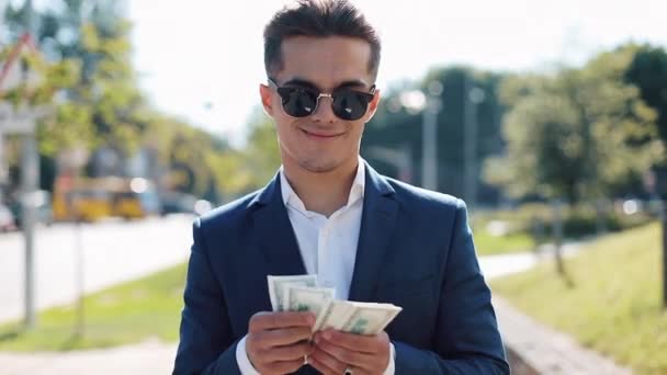 Young happy businesman in sunglasses and a suit counting money and walking in the street. He selebrating his successful win with a lot of dollars - Кадры, видео